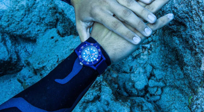 An image of a diver underwater wearing a blue watch. In the background, you can see coral with a blue tinge.