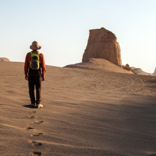 Image of a man walking up a sand dune. The background contains orange and rust colour sand, with a sand rock in the centre.